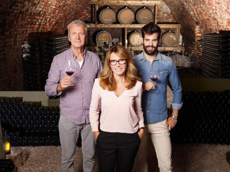 Portrait of multigenerational winery owner family standing at wine cellar. Senior winemaker and young sommelier standing at background and holding in hands a glass of red wine while middle age businesswoman looking at camera and smiling. Small business.